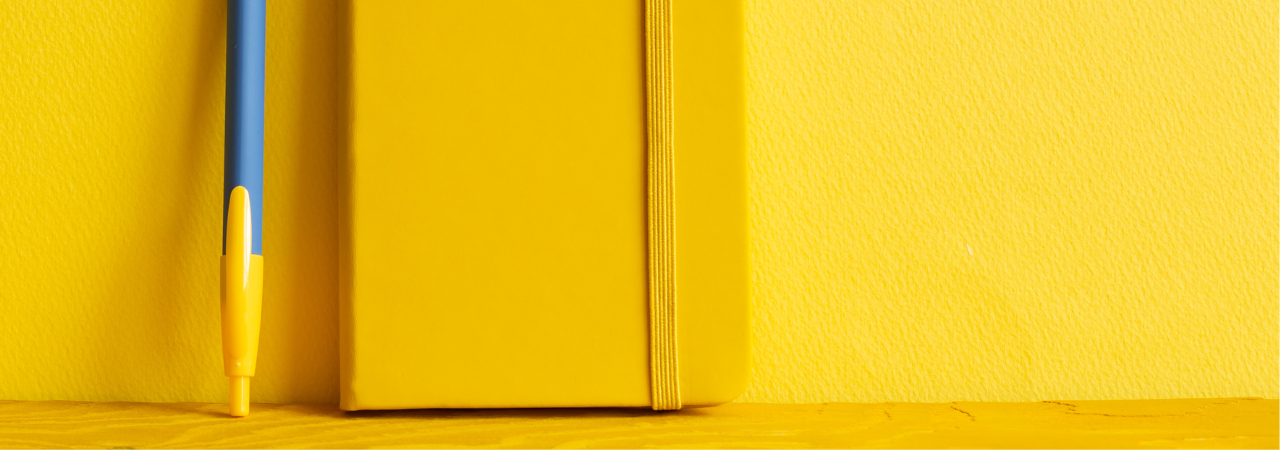 Yellow background with close up focus on yellow notebook and blue and yellow pen