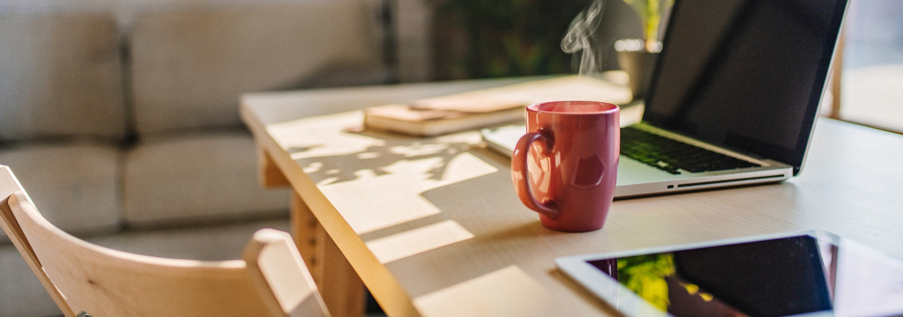 desk with steaming mug in focus, and laptop, tablet, and notebook out of focus in the background