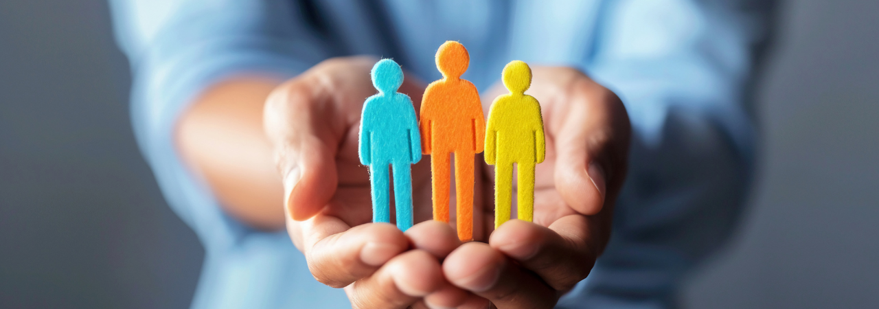 A person wearing a blue shirt holds out their hands, presenting three colorful human-shaped cutouts in blue, orange, and yellow, symbolizing teamwork, diversity, and employee support.